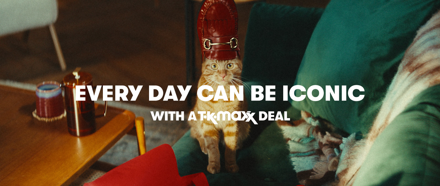 A ginger cat sitting upright on a green sofa with a shiny red loafer on its head in front of a red TK Maxx shopping bag.