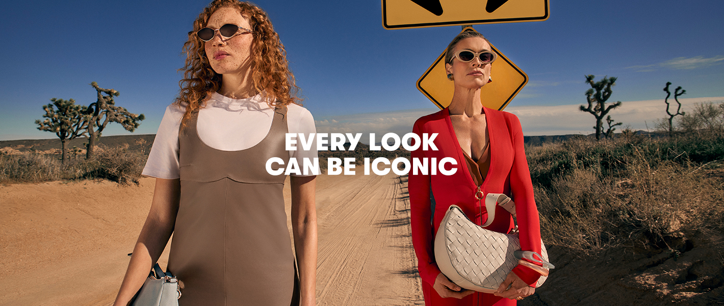 Two people wearing sunglasses stand on a desert road. One wears a T‑shirt layered under a slip dress and holds a handbag. The other wears a red dress and carries a quilted handbag. 