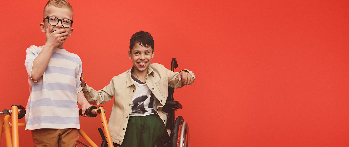 Two boys, one using a walking frame, and one in a wheelchair smile and laugh. The boy with the walking frame has his hand over his mouth.