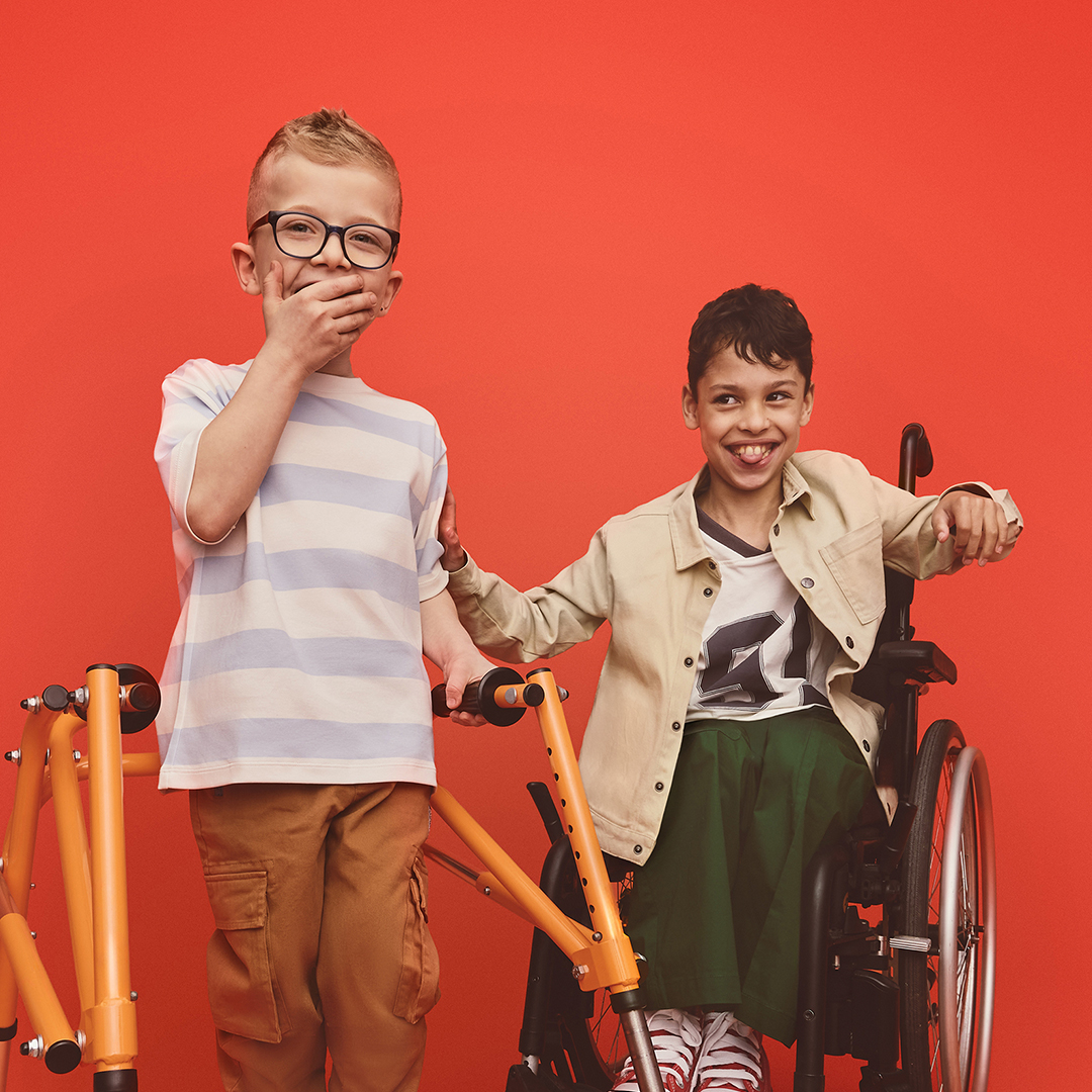 Two boys, one using a walking frame, and one in a wheelchair smile and laugh. The boy with the walking frame has his hand over his mouth.